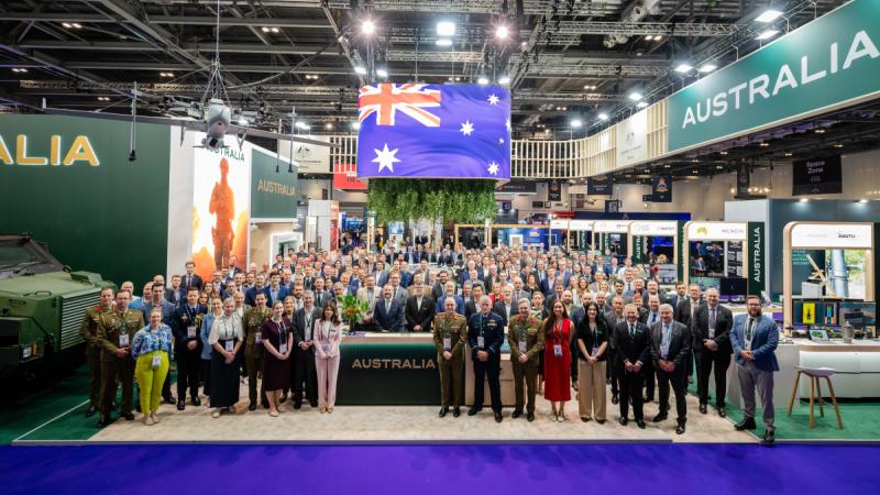 A large group of people gather for a photo in amongst the exhibits at a defence tradeshow.