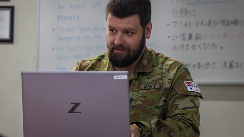 A man in uniform working on a laptop computer, with a whiteboard filled with writing behind him.