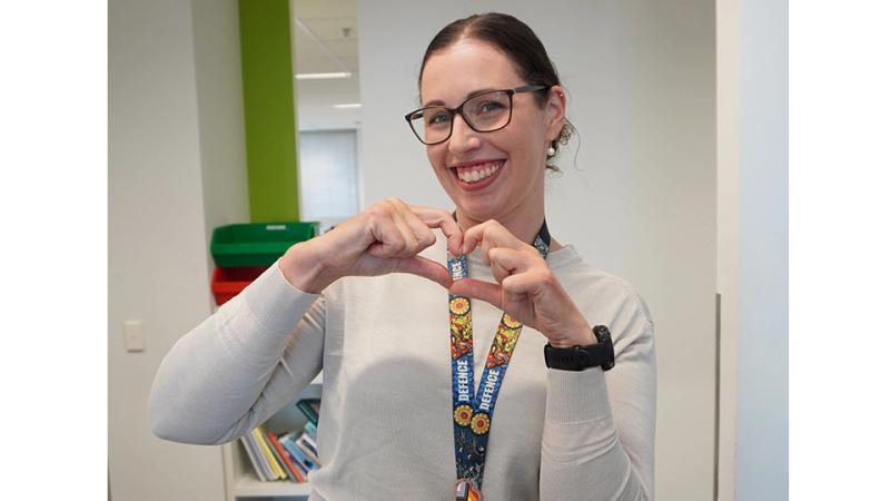 A woman wearing a Defence lanyard smiles and makes a heart shape with her hands while standing in an office hallway.